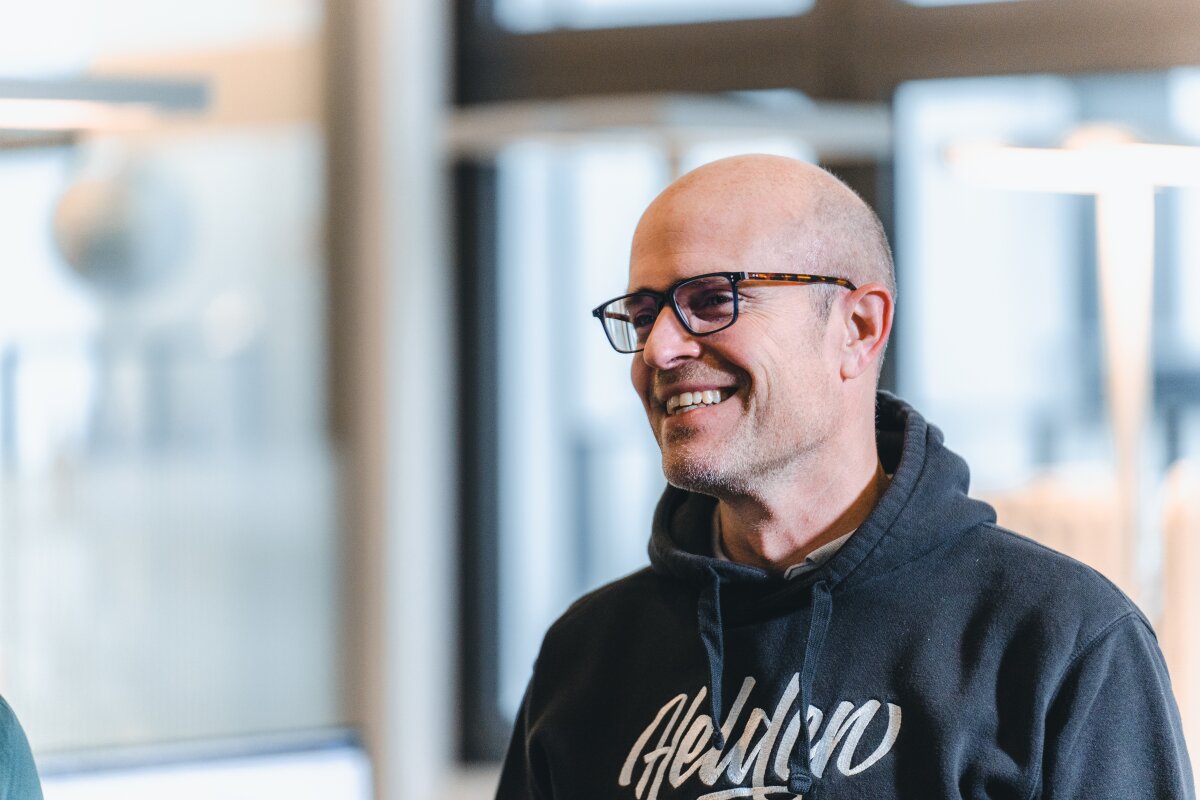 Mitarbeiter der Agentur Homepage Helden im Kapuzenpulli lächelt im hellen Büro.