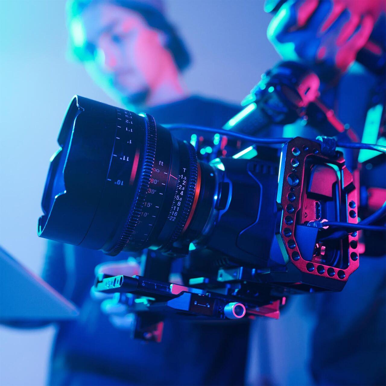 Professionelles Kameraobjektiv an einer Filmkamera, montiert auf einem Rig; Fokusring und Kabel sichtbar, unscharfe Person im Hintergrund, neonblaue und rosa Studiobeleuchtung.