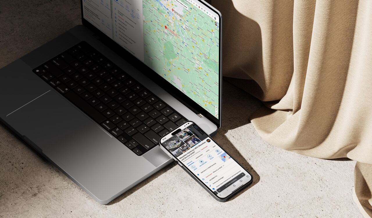 Grauer Laptop mit geöffneter Kartenansicht und ein Smartphone, das eine Geschäftseintragsseite anzeigt; beide liegen auf Betonboden neben beigem, drapiertem Vorhang.