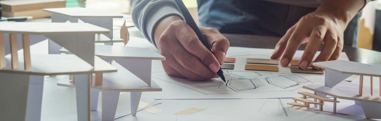 Architekt zeichnet am Arbeitstisch einen Grundriss, daneben Arbeitsmodelle aus weißem Karton und Materialmuster.