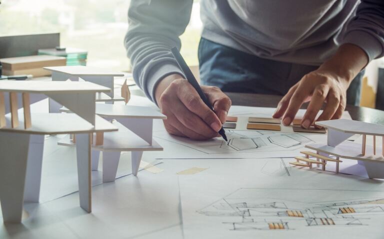 Architekt zeichnet am Arbeitstisch einen Grundriss, daneben Arbeitsmodelle aus weißem Karton und Materialmuster.