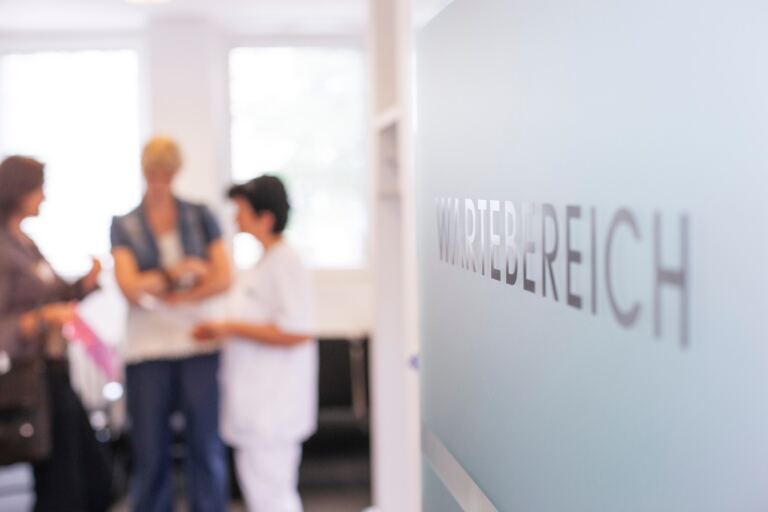 Blick in den Wartebereich einer Arztpraxis, eine Mitarbeiterin im Gespräch mit zwei Patientinnen.