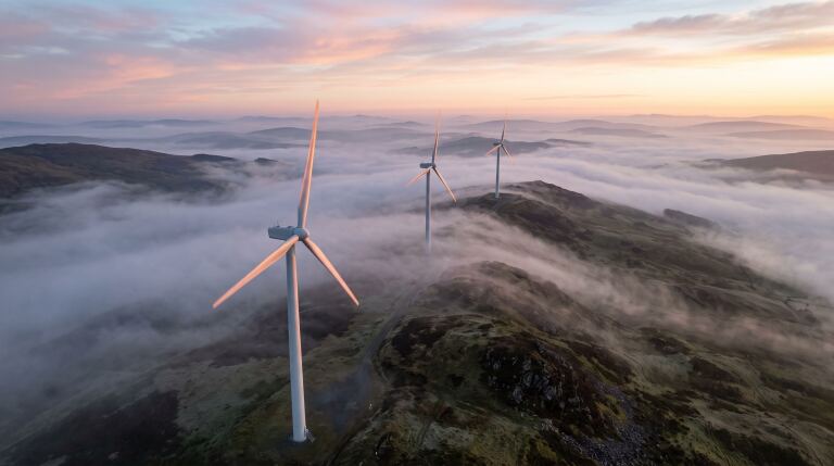 Drei Windenergieanlagen auf einem Höhenzug im Morgenlicht, darunter liegt Nebel in den Tälern.