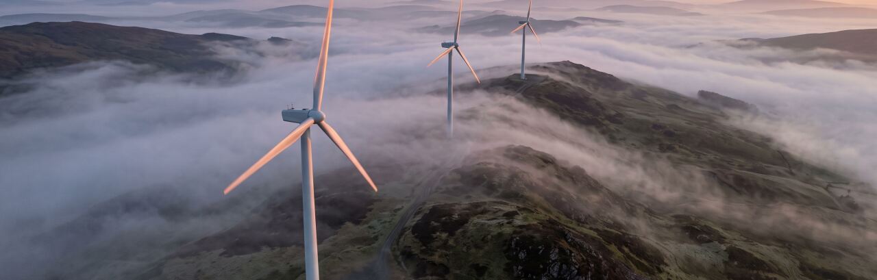 Drei Windenergieanlagen auf einem Höhenzug im Morgenlicht, darunter liegt Nebel in den Tälern.