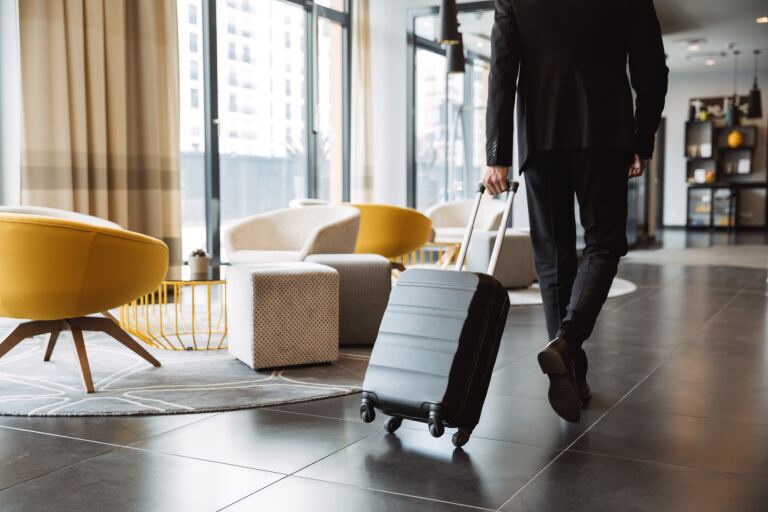 Gast mit Rollkoffer durchquert die Lobby eines modernen Hotels mit gelben Sesseln und Fensterfront.