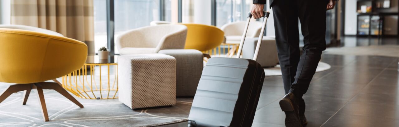 Gast mit Rollkoffer durchquert die Lobby eines modernen Hotels mit gelben Sesseln und Fensterfront.
