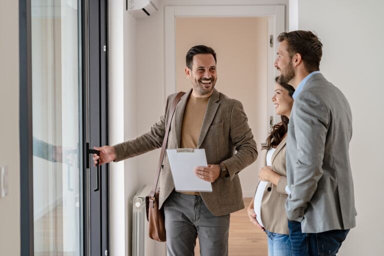 Immobilienmakler öffnet die Terrassentür einer Wohnung und zeigt sie einem jungen Paar.