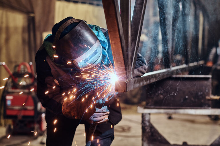 Schweißer mit Schutzschild und Lederhandschuhen setzt in der Werkstatt eine Naht an einer Stahlkonstruktion, Funkenflug.