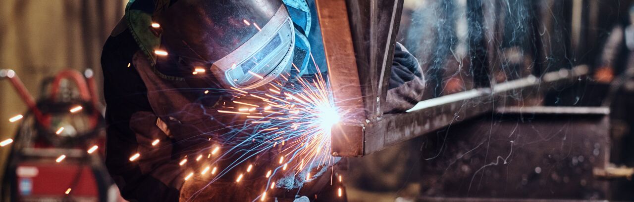 Schweißer mit Schutzschild und Lederhandschuhen setzt in der Werkstatt eine Naht an einer Stahlkonstruktion, Funkenflug.