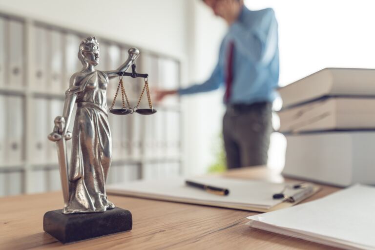Justitia-Figur auf dem Schreibtisch einer Kanzlei, im Hintergrund ein Rechtsanwalt am Aktenregal.
