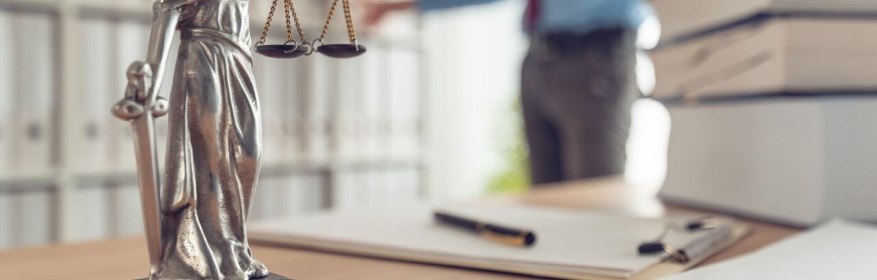 Justitia-Figur auf dem Schreibtisch einer Kanzlei, im Hintergrund ein Rechtsanwalt am Aktenregal.