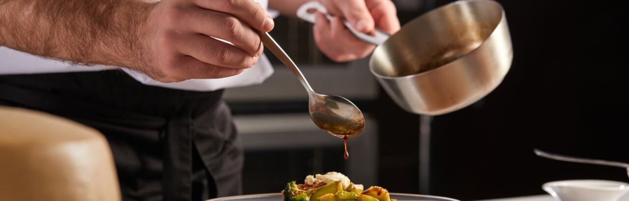 Koch in weißer Jacke nappiert in der Restaurantküche einen Teller mit Brokkoli und Avocado aus einem Löffel mit Sauce.