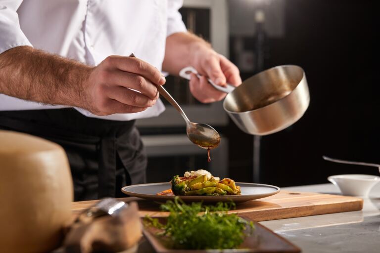 Koch in weißer Jacke nappiert in der Restaurantküche einen Teller mit Brokkoli und Avocado aus einem Löffel mit Sauce.
