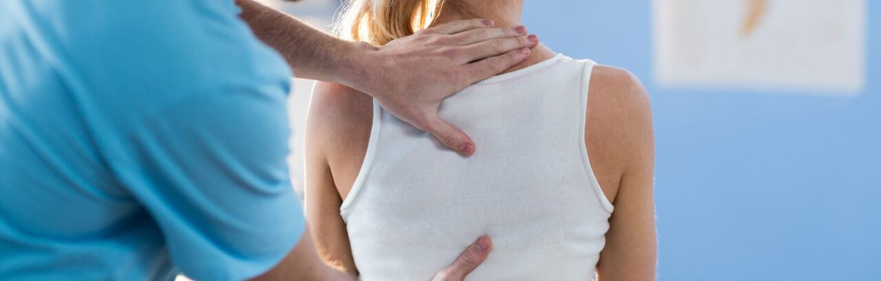Therapeut tastet mit beiden Händen den Rücken einer Patientin ab, im Hintergrund eine Schautafel der Wirbelsäule.