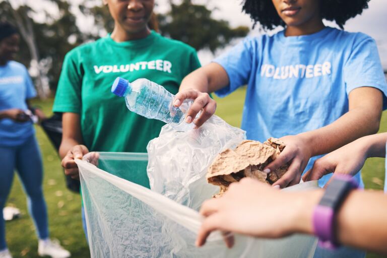 Freiwillige in Vereins-T-Shirts sammeln bei einer Aufräumaktion im Park Müll in einen Sack: eine Plastikflasche und zerknülltes Papier werden eingeworfen.