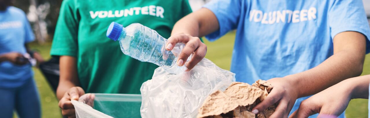 Freiwillige in Vereins-T-Shirts sammeln bei einer Aufräumaktion im Park Müll in einen Sack: eine Plastikflasche und zerknülltes Papier werden eingeworfen.