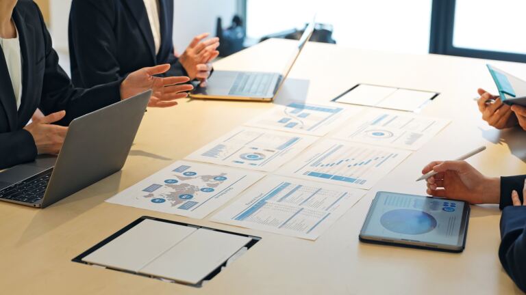 Besprechung am Konferenztisch: ausgebreitete Auswertungen mit Diagrammen, Laptops und ein Tablet mit Kennzahlen.