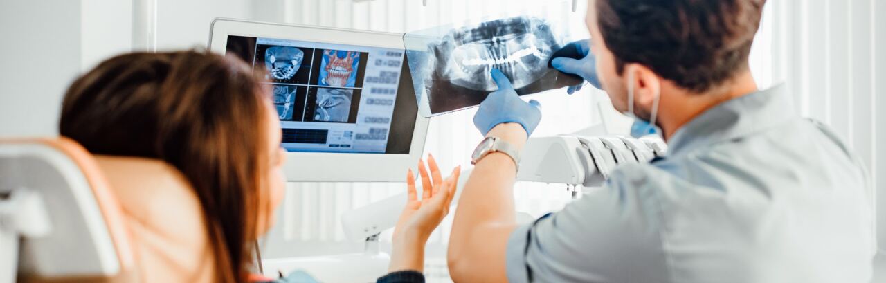 Zahnarzt erklärt einer Patientin im Behandlungsstuhl ein Röntgenbild des Kiefers, im Hintergrund der Bildschirm mit der Aufnahme.