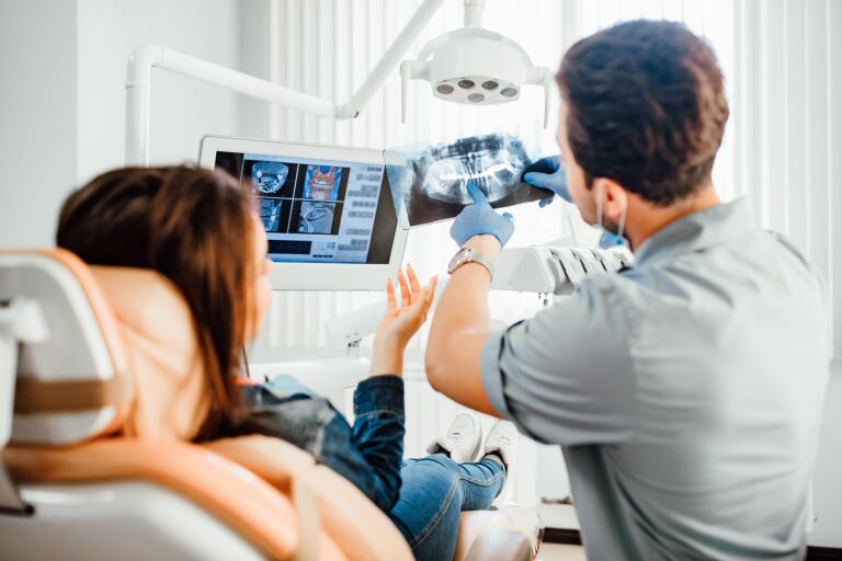 Zahnarzt erklärt einer Patientin im Behandlungsstuhl ein Röntgenbild des Kiefers, im Hintergrund der Bildschirm mit der Aufnahme.