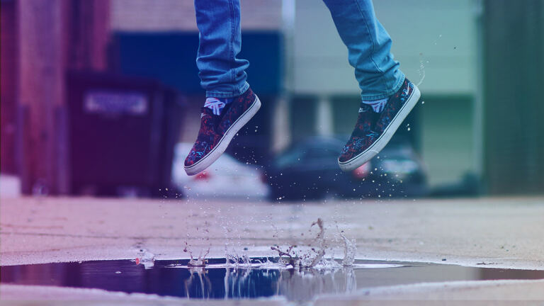 Untere Beine in blauen Jeans und gemusterten Slip-On-Sneakers springen über eine Pfütze; Wasser spritzt hoch, städtischer Bürgersteig mit unscharfen Gebäuden und parkenden Autos im Hintergrund.