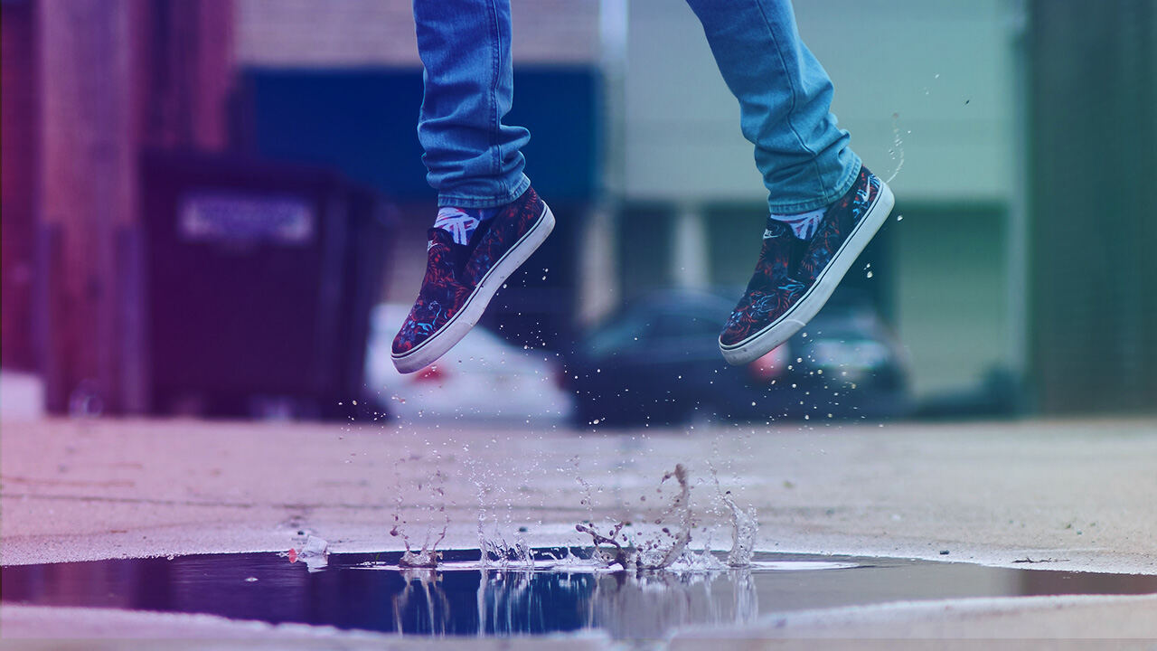 Untere Beine in blauen Jeans und gemusterten Slip-On-Sneakers springen über eine Pfütze; Wasser spritzt hoch, städtischer Bürgersteig mit unscharfen Gebäuden und parkenden Autos im Hintergrund.