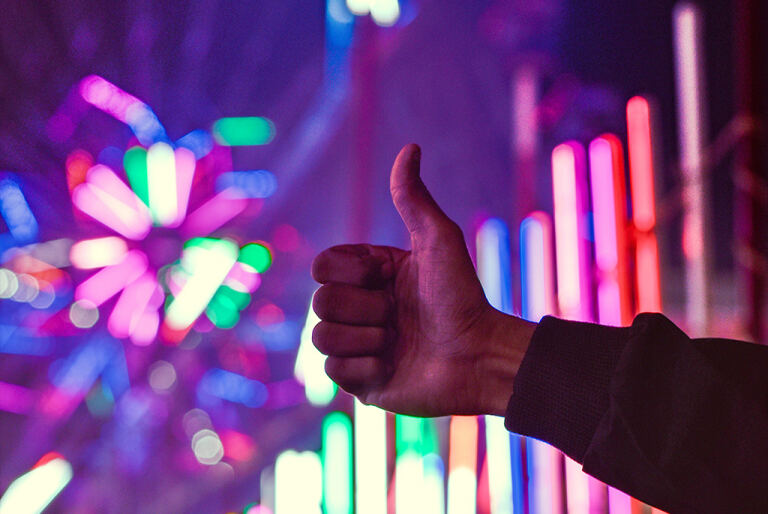 Hand mit nach oben gestrecktem Daumen – Daumen hoch – vor unscharfen, bunten Neonlichtern und einem verschwommenen Riesenrad in lila, pink, grün und blau bei Nacht.