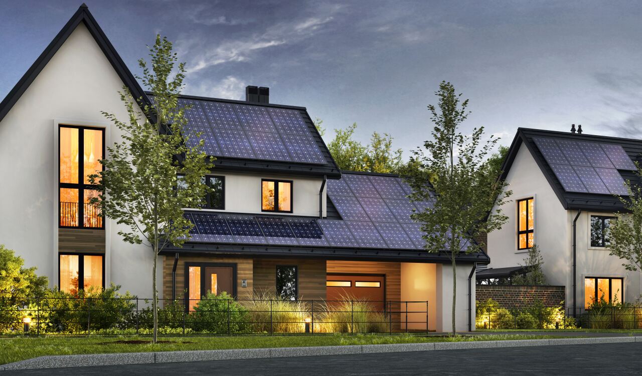 Zweistöckiges Wohnhaus mit Solarpaneelen auf den Dächern – beleuchtete Fenster innen – Straßenfront mit Garage, eingezäuntem Vorgarten, Sträuchern, Bäumen und wolkigem Abendhimmel.