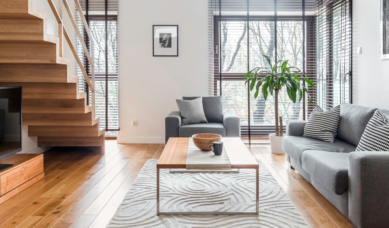 Graues Sofa und grauer Sessel stehen gegenüber einem hölzernen Couchtisch, in hellem Wohnzimmer mit Treppe links, großen Fenstern mit Jalousien, Holzfußboden und Zimmerpflanze.