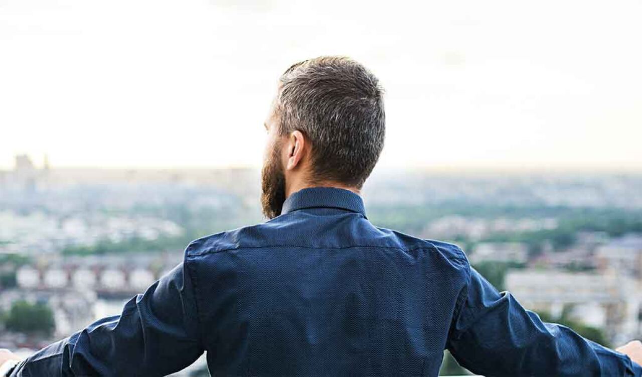Mann mit kurzem Haar und Bart – steht mit ausgebreiteten Armen auf einer Brüstung – Blick über unscharfe Stadtsilhouette mit Wohnhäusern und Grünflächen bei Tageslicht.