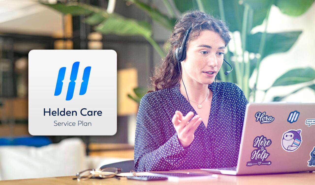 Frau mit Headset gestikuliert vor Laptop; links Logo 'Helden Care' 'Service Plan'; Laptopsticker mit Texten 'Hero', 'Here to Help', 'SEND HELP'; Büro mit Pflanzen im Hintergrund.