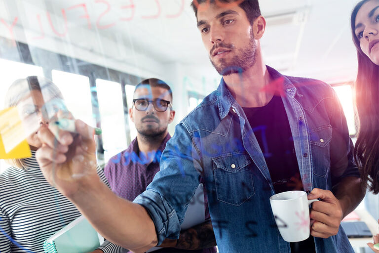 Junger Mann schreibt mit Marker auf eine Glaswand und hält eine Kaffeetasse; drei Kollegen beobachten, Post‑it‑Zettel und reflektierende Markerbeschriftungen auf dem Glas, heller Büroraum